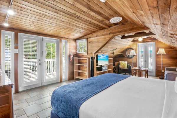 A cozy wooden bedroom with a large bed, TV, seating area, and doors opening to a balcony. Natural light fills the space.