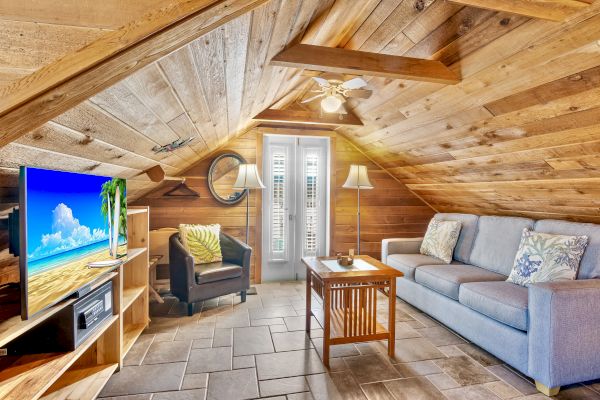 A cozy attic room with wooden walls, a TV, sofa, armchair, coffee table, and a ceiling fan, featuring a bright window.