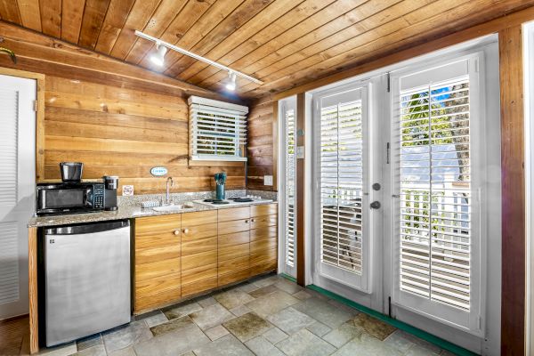 A cozy kitchen with wooden walls features a small fridge, sink, coffee maker, and microwave, leading to a patio through French doors.