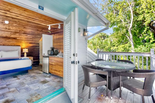 A cozy room with wood accents opens to a balcony featuring a round table and chairs, surrounded by lush greenery in the background.