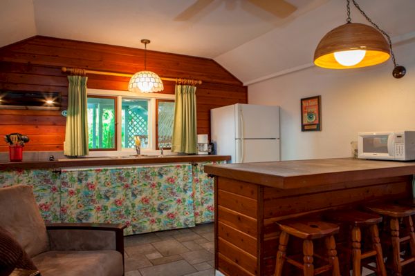 A cozy kitchen with wooden accents, a floral curtain, bar stools, a microwave, and hanging lamps, creating a warm and inviting atmosphere.