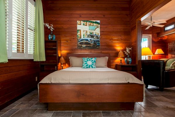 A cozy bedroom with a wooden finish, a large bed, vibrant pillow, artwork above, and ambient lighting from lamps.