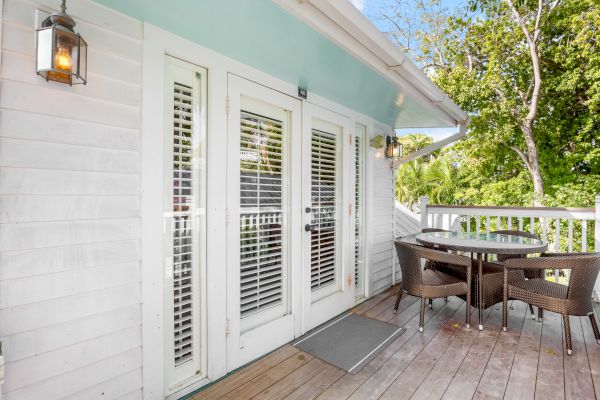 A wooden deck with a small glass table and chairs is alongside double doors with blinds, surrounded by greenery and a wall lantern.