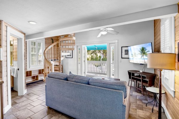 A cozy living room with a spiral staircase, sofa, TV, and bright decor. French doors lead to a porch with palm tree view outside.