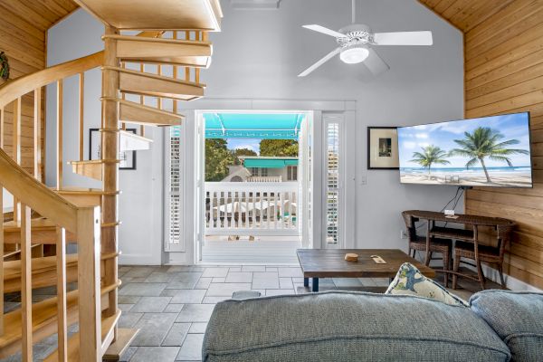 The image shows a cozy living room with a spiral staircase, a ceiling fan, TV, and a view of a balcony with palm trees outside.