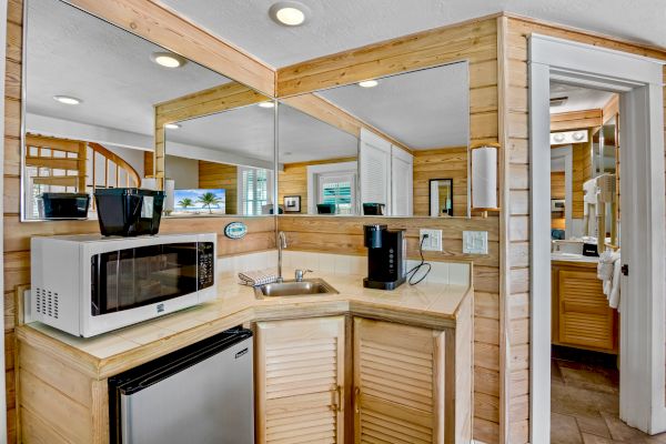 A small kitchenette with a microwave, mini-fridge, coffee maker, sink, and wooden cabinets next to a mirrored wall and a door.