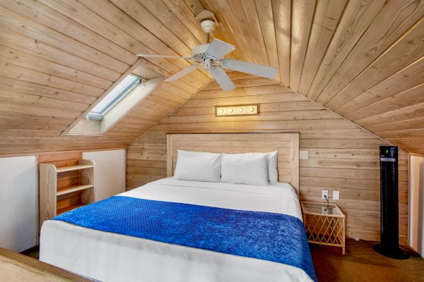 A cozy attic bedroom with wooden ceiling, double bed, blue blanket, ceiling fan, skylight, small shelf, and side table with a fan.