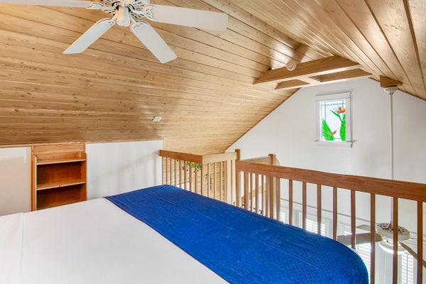 The image shows a cozy loft bedroom with a wooden ceiling, white walls, a bed with a blue blanket, a ceiling fan, and a small window.