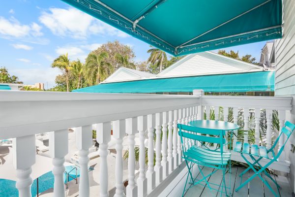 The image shows a balcony with turquoise chairs and table, a white railing, awning, and a view of rooftops and palm trees on a sunny day.