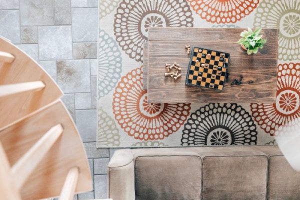 A cozy room with a patterned rug, a wooden coffee table with a chessboard, some chess pieces, a plant, and a beige couch next to a spiral staircase.