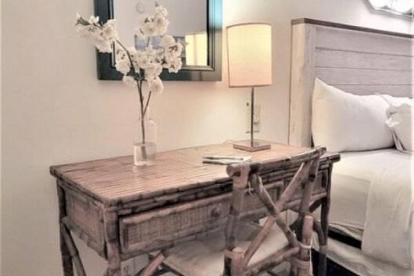 The image shows a cozy bedroom corner with a wooden desk, a chair, a vase with flowers, a lamp, and part of a bed in a minimal style.