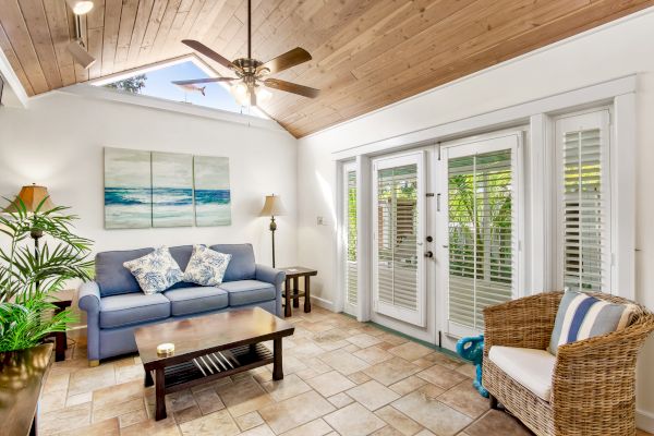 A cozy living room with a blue sofa, coffee table, wicker chair, and ceiling fan. Glass doors lead outside. Art decorates the wall.