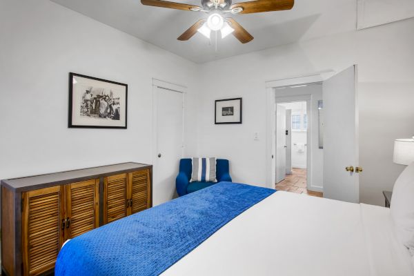 A bedroom with a blue bedspread, wooden cabinet, artwork on the walls, and a ceiling fan. There's also a blue chair near the corner.