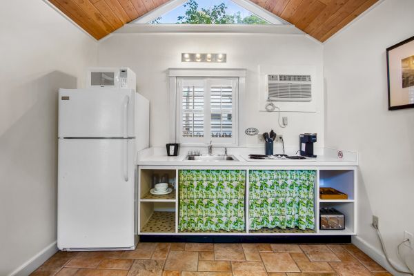 A small kitchenette with a fridge, microwave, sink, and shelves with floral curtains. Coffee makers and a window are above the sink.