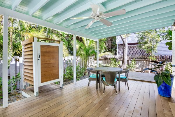 The image shows a covered outdoor patio with a ceiling fan, table and chairs, wooden flooring, and surrounding greenery in a backyard setting.