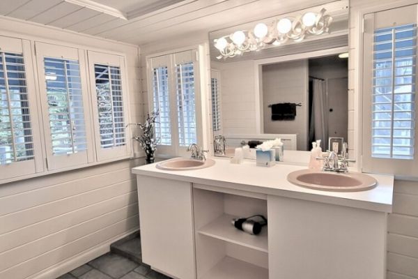 The image shows a bathroom with two sinks, mirrors, bright lights, and windows with shutters. The decor is white and neat.