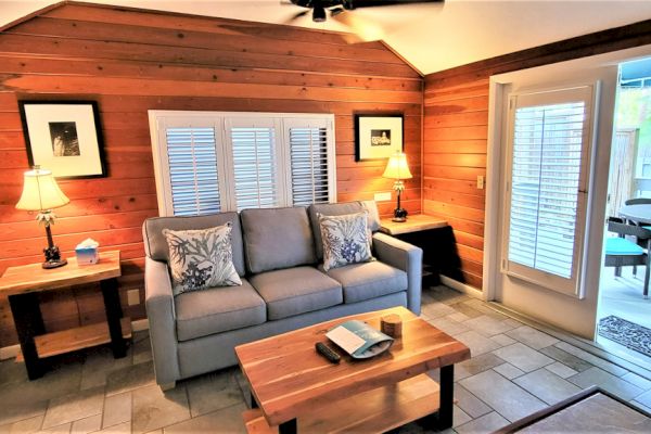 A cozy living room with a gray sofa, two lamps, wooden walls, coffee table, and a view of the patio through glass doors.