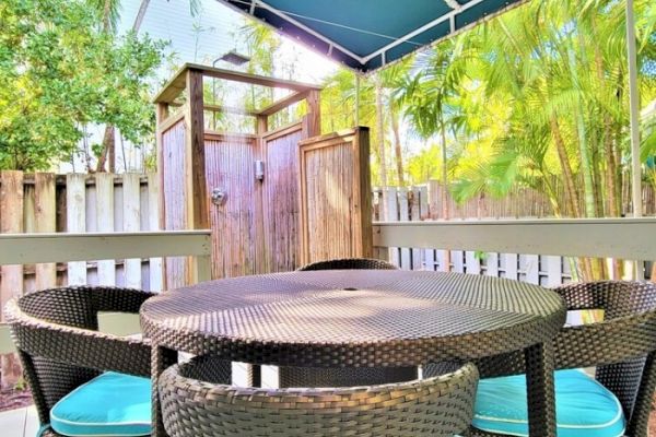 A patio with a round wicker table and chairs under a canopy, surrounded by greenery and featuring a wooden outdoor shower enclosure.