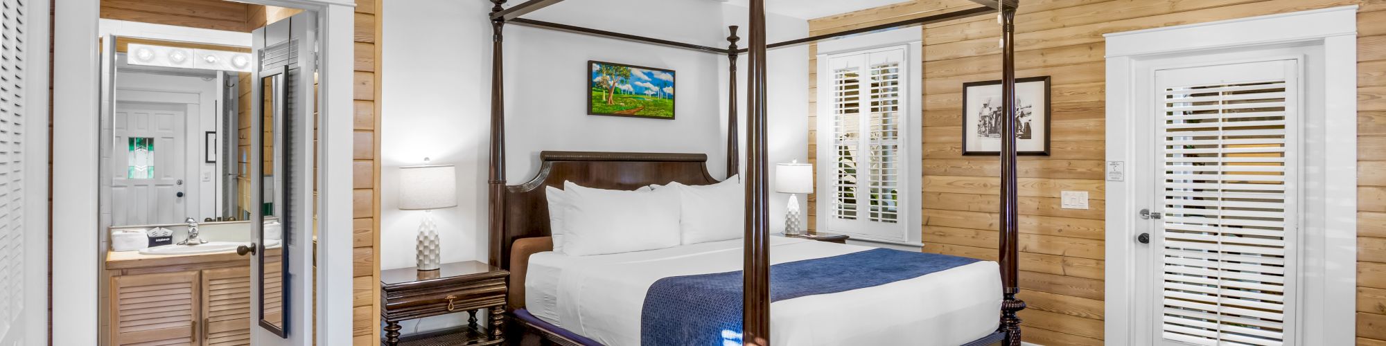 A cozy bedroom with a four-poster bed, wooden walls, a ceiling fan, and a neat bathroom view. Blinds on the door let in natural light.