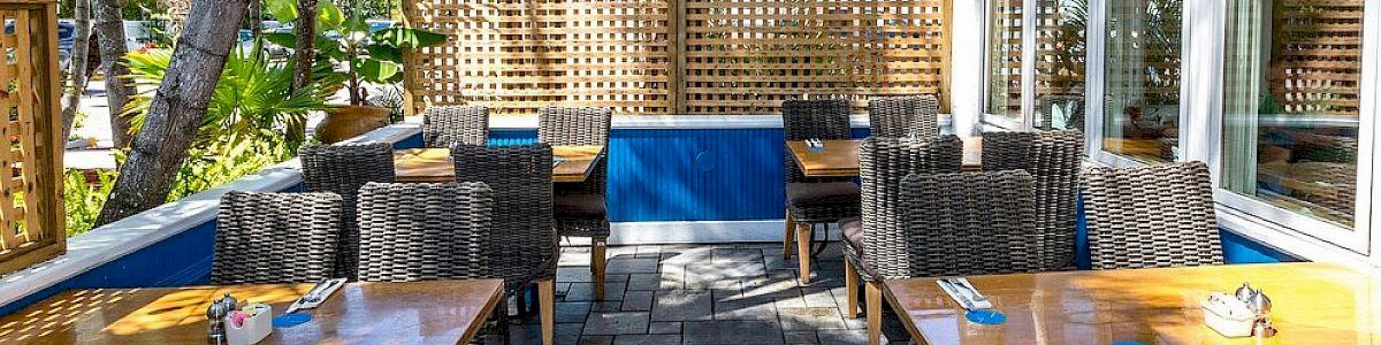 Outdoor patio dining area with wooden tables, wicker chairs, and lush greenery. Sunny and inviting atmosphere.