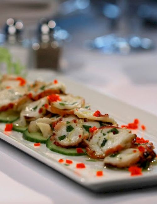 Thinly sliced octopus served on a rectangular plate, garnished with herbs and diced red peppers, with lettuce on the side.