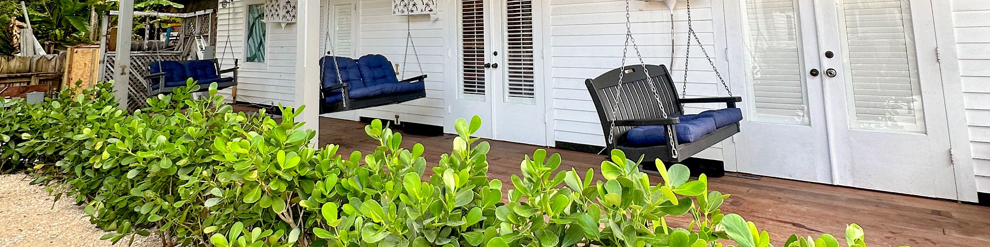 A white two-story house with green awnings, a large porch, swings, and plants in the foreground.