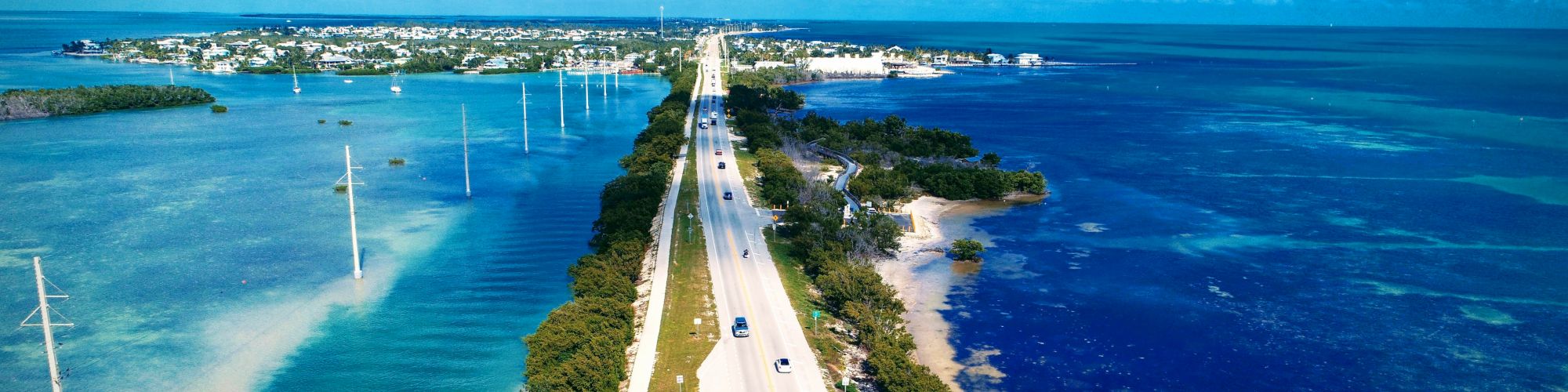 Aerial view of a highway flanked by blue waters and trees, leading to a distant town under a partly cloudy sky.