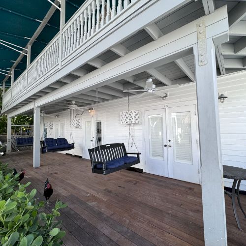 A patio with swings, a table, and chairs under a covered porch; greenery surrounds and a rooster is strolling nearby.