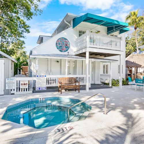 A small outdoor pool in front of a white two-story house with a balcony and patio furniture, surrounded by trees and clear blue sky.