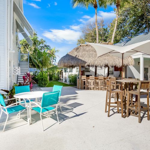 Outdoor patio with a table, chairs, tiki bar, and umbrella; surrounded by tropical trees and a building.