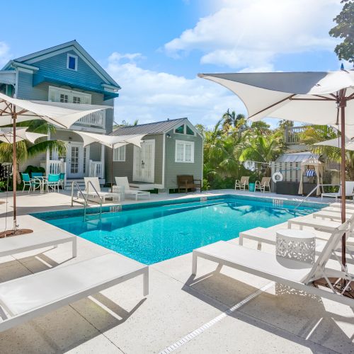 The image shows a luxurious poolside area with loungers and umbrellas, set in front of a stylish house with palm trees and a blue sky.