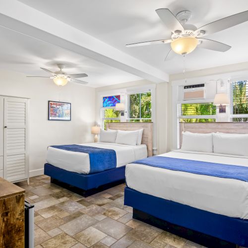 The image shows a bright room with two double beds, ceiling fans, a wooden dresser, and windows with shutters and natural light.