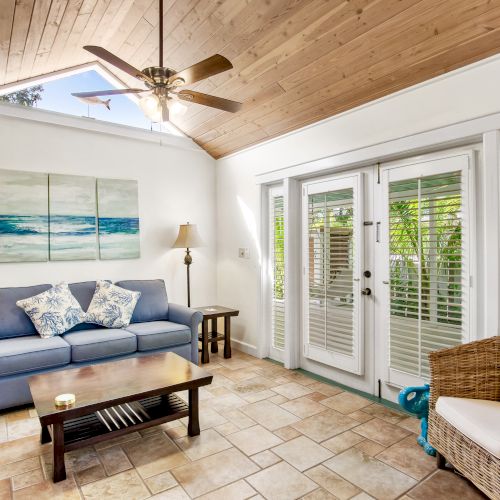 A cozy living room features a blue sofa, rattan chair, wooden coffee table, ceiling fan, large windows, and ocean-themed artwork on the wall.