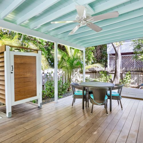 Covered patio with a ceiling fan, wooden flooring, outdoor shower, round table, chairs, plants, and garden view, creating a relaxing vibe.