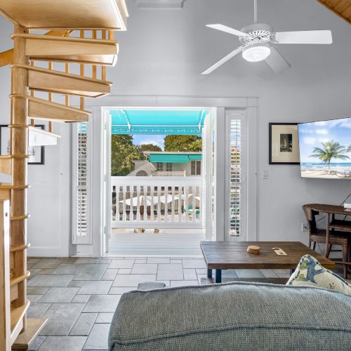 A cozy living room with a spiral staircase, sofa, TV showing a beach scene, and a view of a balcony with a canopy.