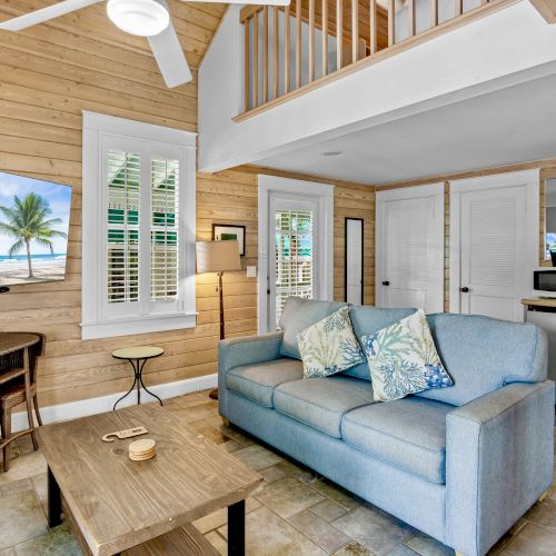 Cozy living area with wooden walls, sofa, TV showing a beach scene, small dining table, and kitchenette, featuring a loft and ceiling fan.