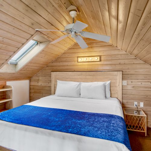 An attic bedroom with wooden walls, a double bed, blue blanket, ceiling fan, skylight, and bedside table with a modern fan.