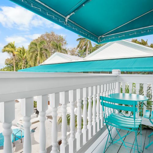 A small balcony with a turquoise table and chairs under a teal awning, overlooking a tropical setting with palm trees and rooftops.