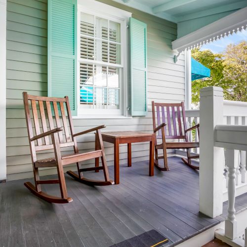 A cozy porch with two wooden rocking chairs and a small table, surrounded by a white railing and light green shutters.