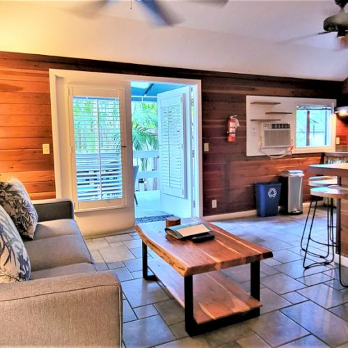 A cozy living area with a sofa, wooden coffee table, bar stools, and kitchen, featuring wooden walls and a door to an outdoor space.