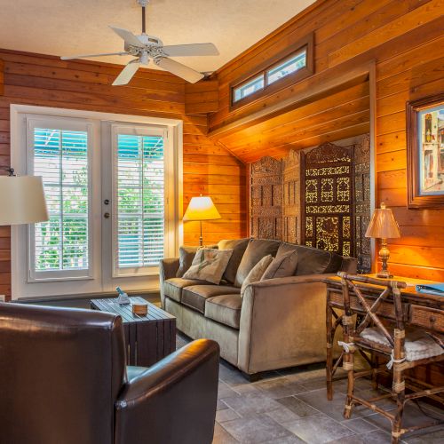 A cozy room with wooden walls, a couch, chairs, lamps, artwork, and a window with blinds. A ceiling fan is installed above the seating area.