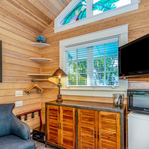 A cozy room with wooden walls, an armchair, shelves, a lamp, cabinets, a TV, microwave, and small fridge under a window with stained glass.