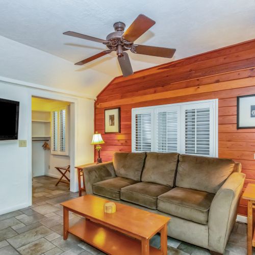 A cozy living room with a brown sofa, two lamps, a ceiling fan, and a wall-mounted TV. The walls have wood paneling for a warm feel.