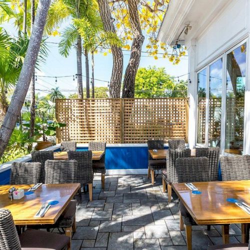 An outdoor patio with tables and wicker chairs set for dining, surrounded by tropical plants and trees with string lights overhead.