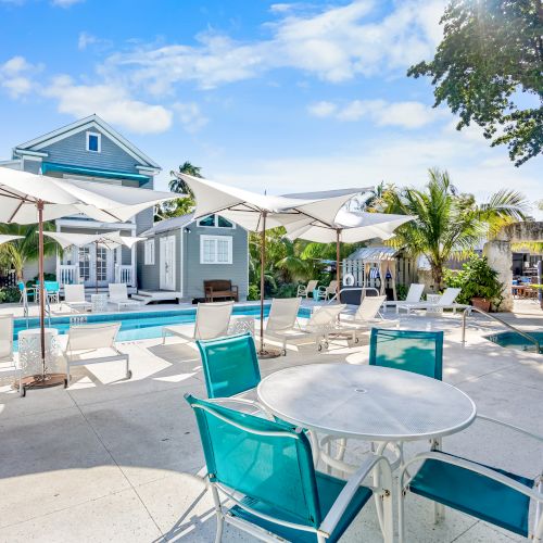 Poolside area with white umbrellas, loungers, and teal chairs around a table. A building and trees are visible in the background.