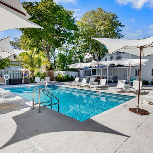 The image shows a sunny pool area with lounge chairs, large umbrellas, and surrounding trees. The setting appears relaxing and inviting.