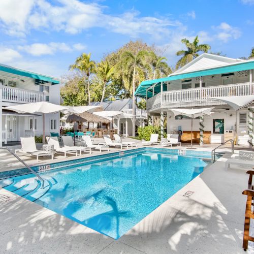 The image shows a sunny courtyard with a swimming pool, surrounded by lounge chairs and white buildings with teal accents and palm trees.