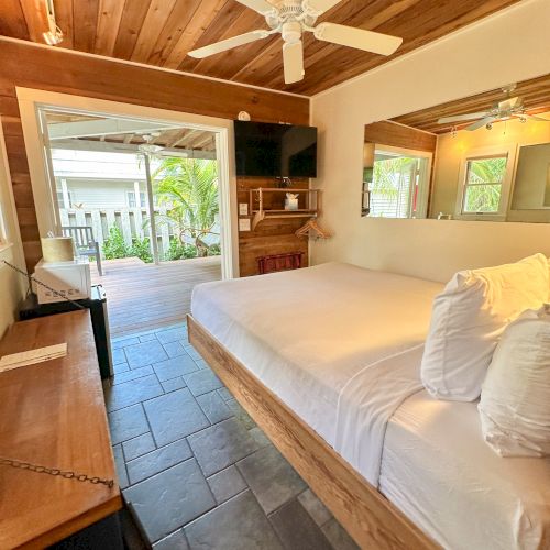 A cozy bedroom with a wooden ceiling, a bed with white linens, a TV, mirror, and open doors leading to an outdoor area with greenery.