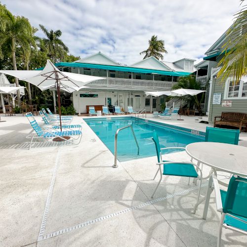 A poolside area with lounge chairs, umbrellas, and a turquoise pool, surrounded by palm trees and buildings under a partly cloudy sky.