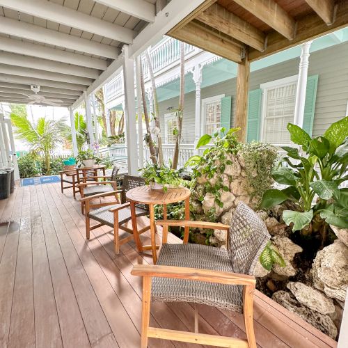 A wooden deck with chairs and tables, surrounded by greenery and partially enclosed by a white railing, in an inviting outdoor setting.